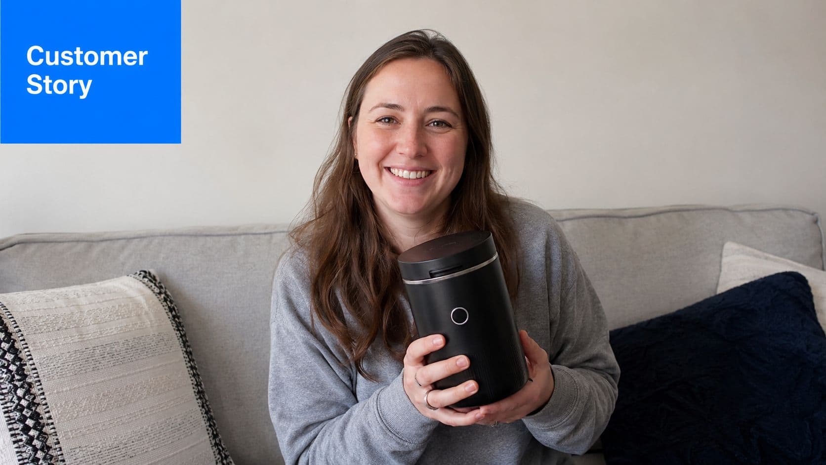 A smiling young woman sits on a sofa holding a black cylinder appliance as a customer story.