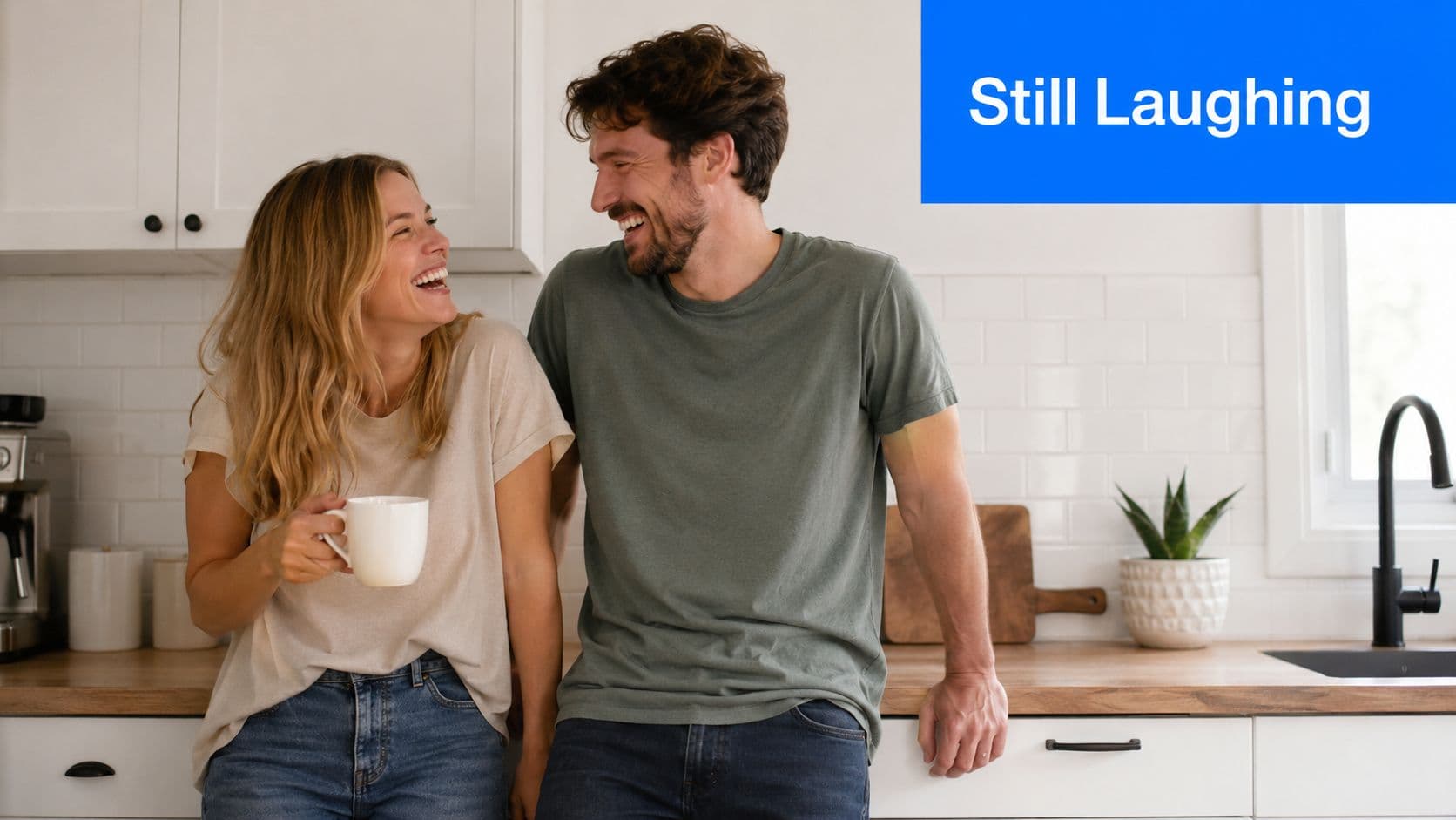 A happy couple laughing together in their kitchen while the woman holds a white coffee mug.